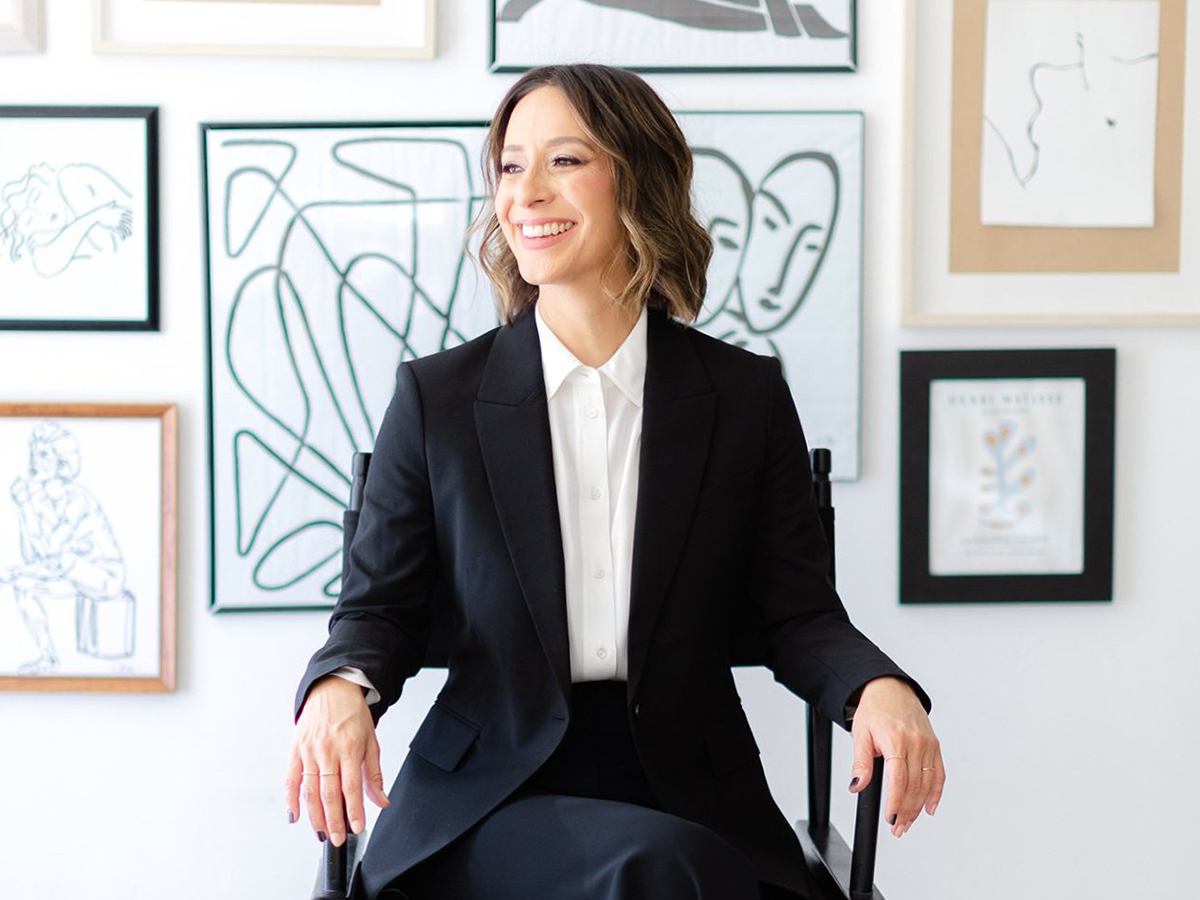 A 3/4 shot of Olga Brandeis sitting in a director's chair in front of a gallery wall with black and white, framed, drawings.